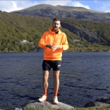 A bearded man in running gear stood on a rock in front of a lake.